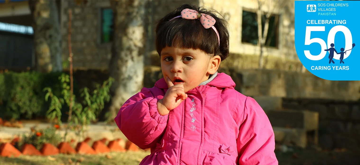 Happy girl in floral dress sits on grass, representing joy at SOS Shelter Homes in Pakistan. Colorful border with icons below.